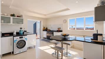 A modern kitchen with a washing machine, a dining area with a table and chairs, and a view of the sea through large windows.