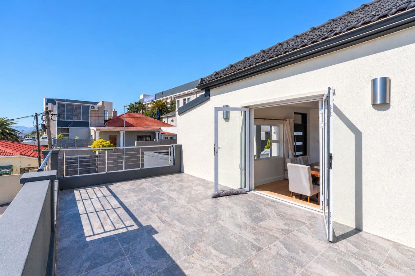 A residential building with a patio area, a sliding glass door leading to an interior space with a table and chairs, and a tiled floor.