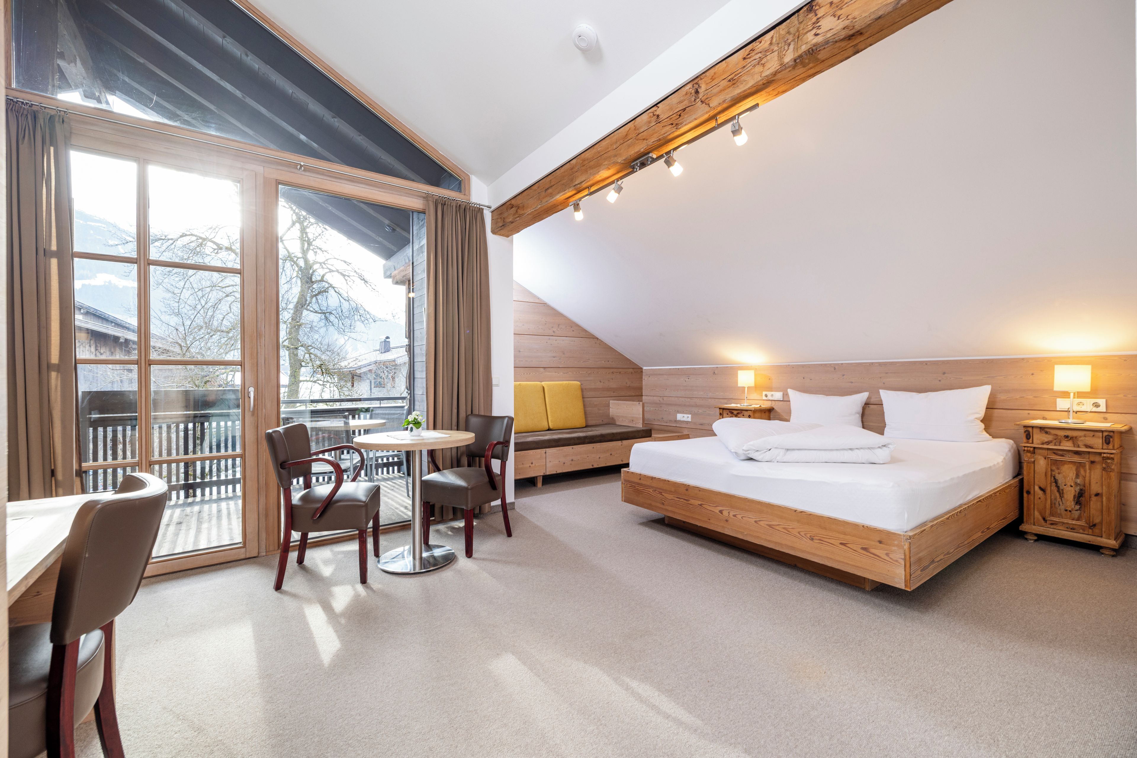 A bedroom with a large bed, a desk, a chair, and a balcony with a view of trees and mountains.