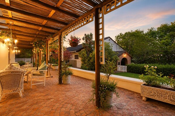 Un patio al aire libre con mobiliario de mimbre, una pérgola y un sendero de ladrillo que conduce a una casa con chimenea de ladrillo.