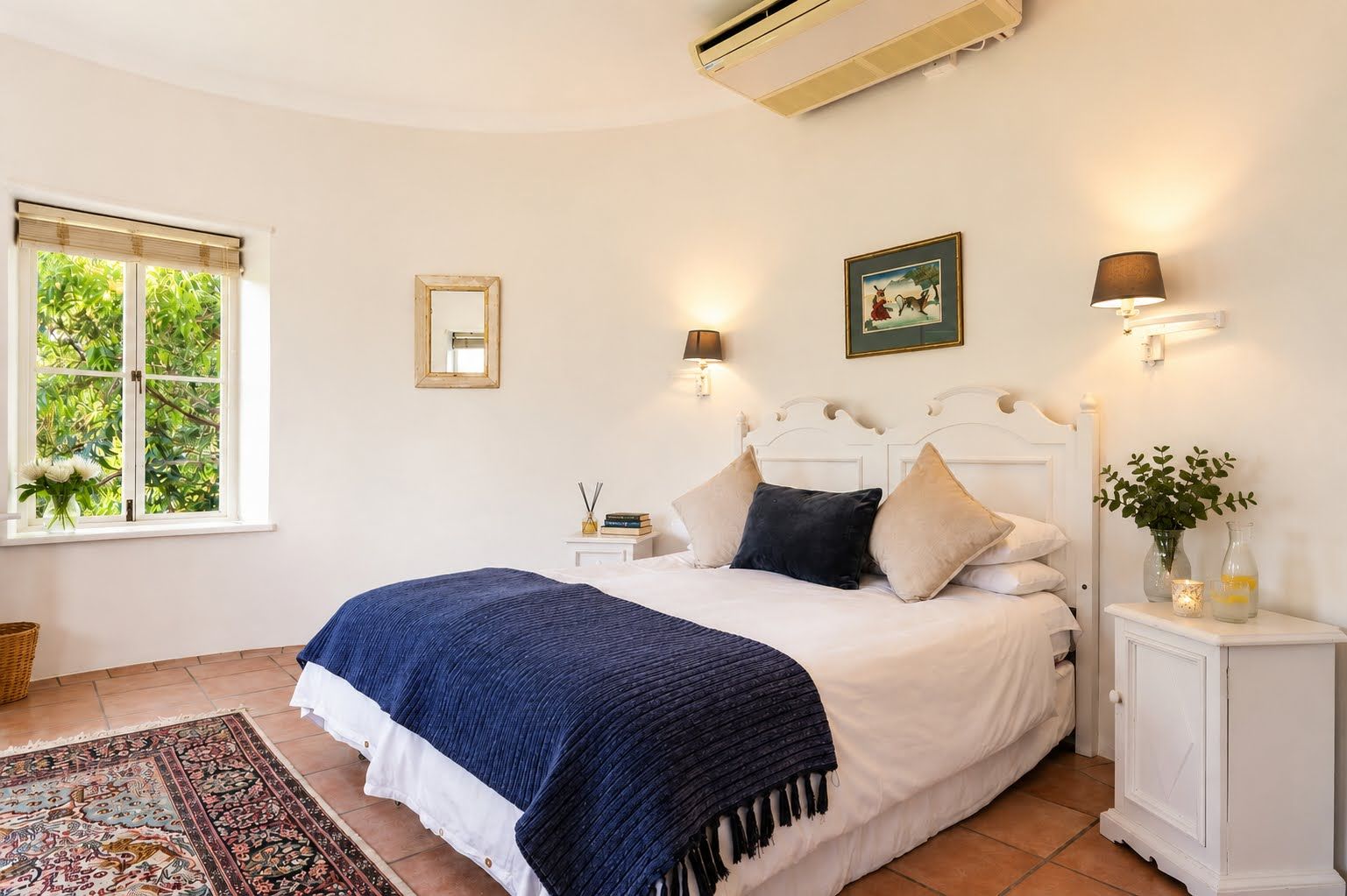 A bedroom with a bed, bedside tables, a nightstand, a window with a view, and an air conditioning unit.