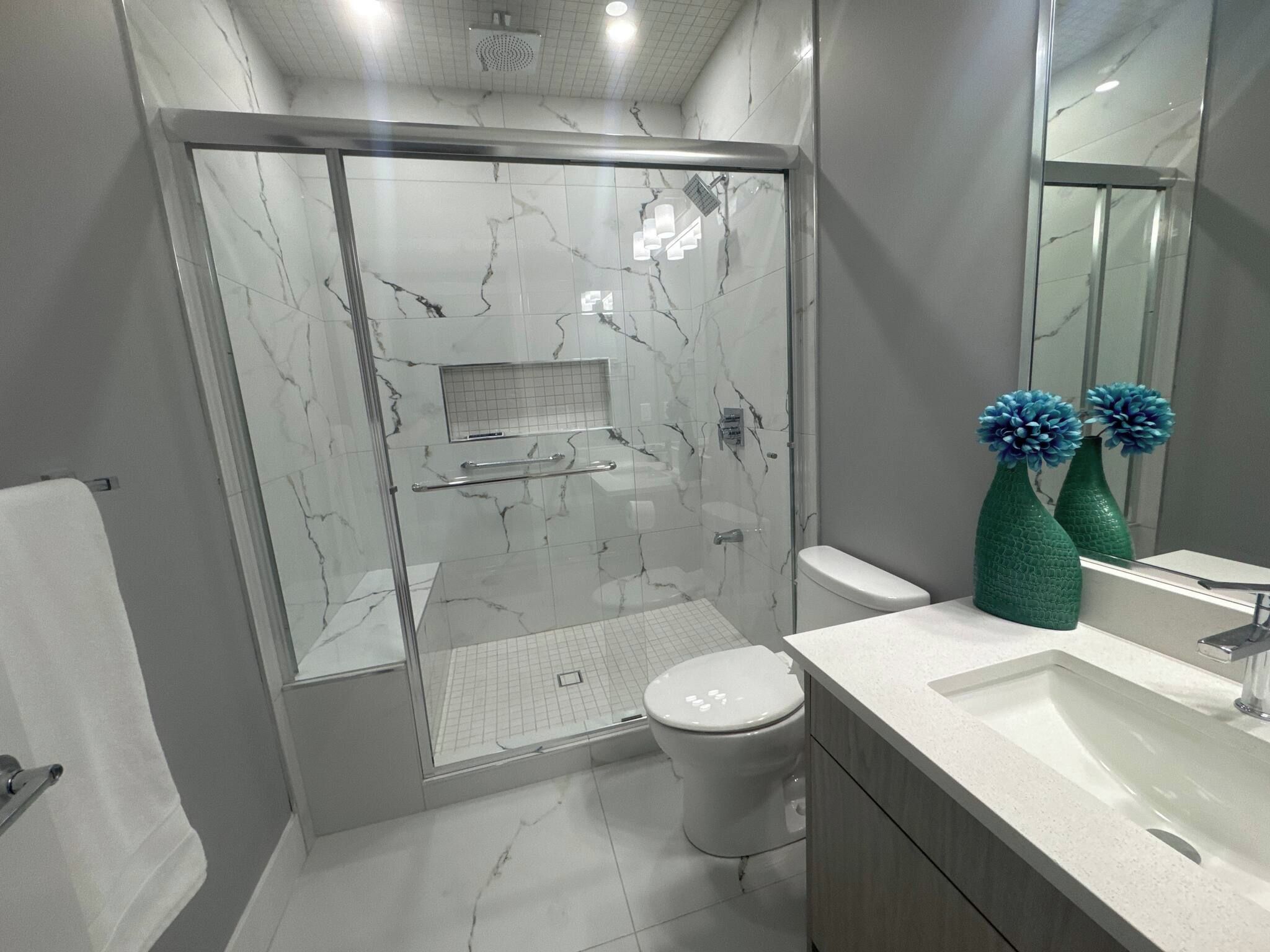 A modern bathroom with a glass-enclosed shower, a white toilet, and a vanity with a mirror and two green vases.