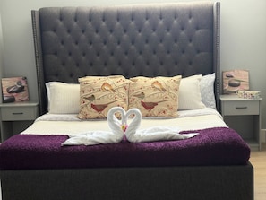 Een bed met een gestoffeerd hoofdbord, twee sierkussens met vogelmotieven en twee handdoeken gevouwen in de vorm van een hart op een paarse deken.