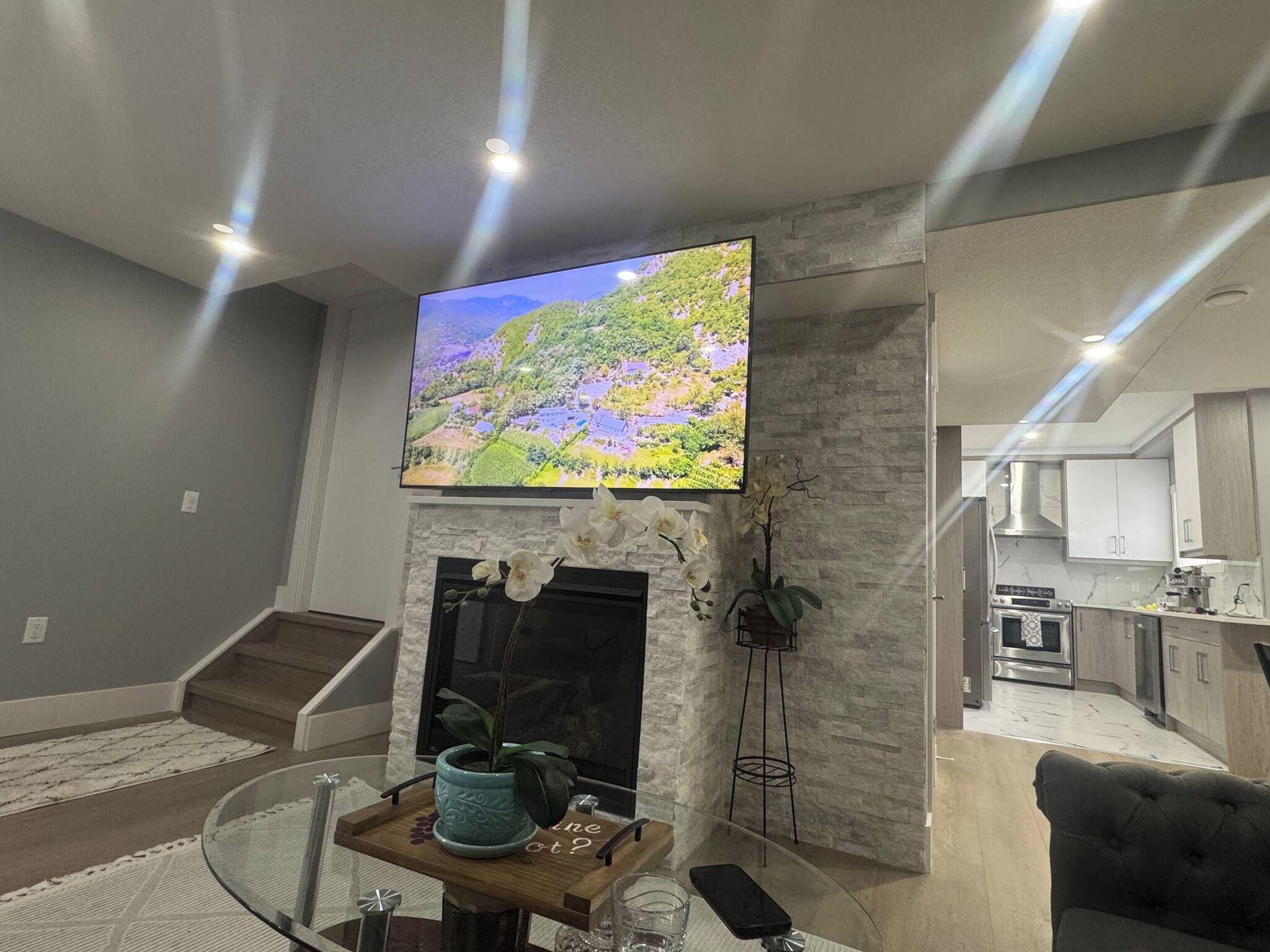 A modern living room with a flat-screen TV mounted on a stone wall, a fireplace, and a kitchen area visible through an open door.