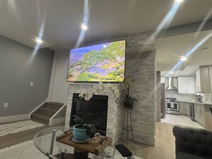 Una sala de estar moderna con un televisor de pantalla plana montado en una pared de piedra, una chimenea y una cocina visible a través de un ventanal.