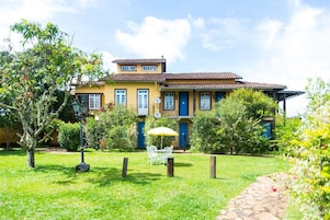 Un edificio amarillo de dos pisos con techo de tejas, rodeado de un jardín bien cuidado con césped, un camino empedrado y una mesa exterior blanca con sillas.