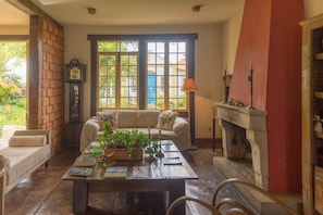Una sala de estar acogedora con chimenea, un sofá, una mesa de centro con libros y plantas, y ventanas grandes con vistas a la vegetación.