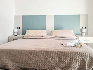 Una cama bien tendida con cubrecama acolchado, dos mesitas de noche con lámparas, y una pared con diseño de panel vertical.