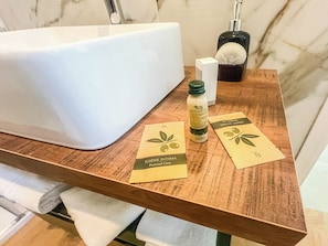 Un tocador de baño con un lavabo blanco, una botella de jabón líquido y dos tarjetas con motivos de hojas de olivo.