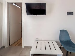 Una habitación moderna con televisor de pantalla plana en la pared, una mesa blanca con toma de corriente y una silla azul.
