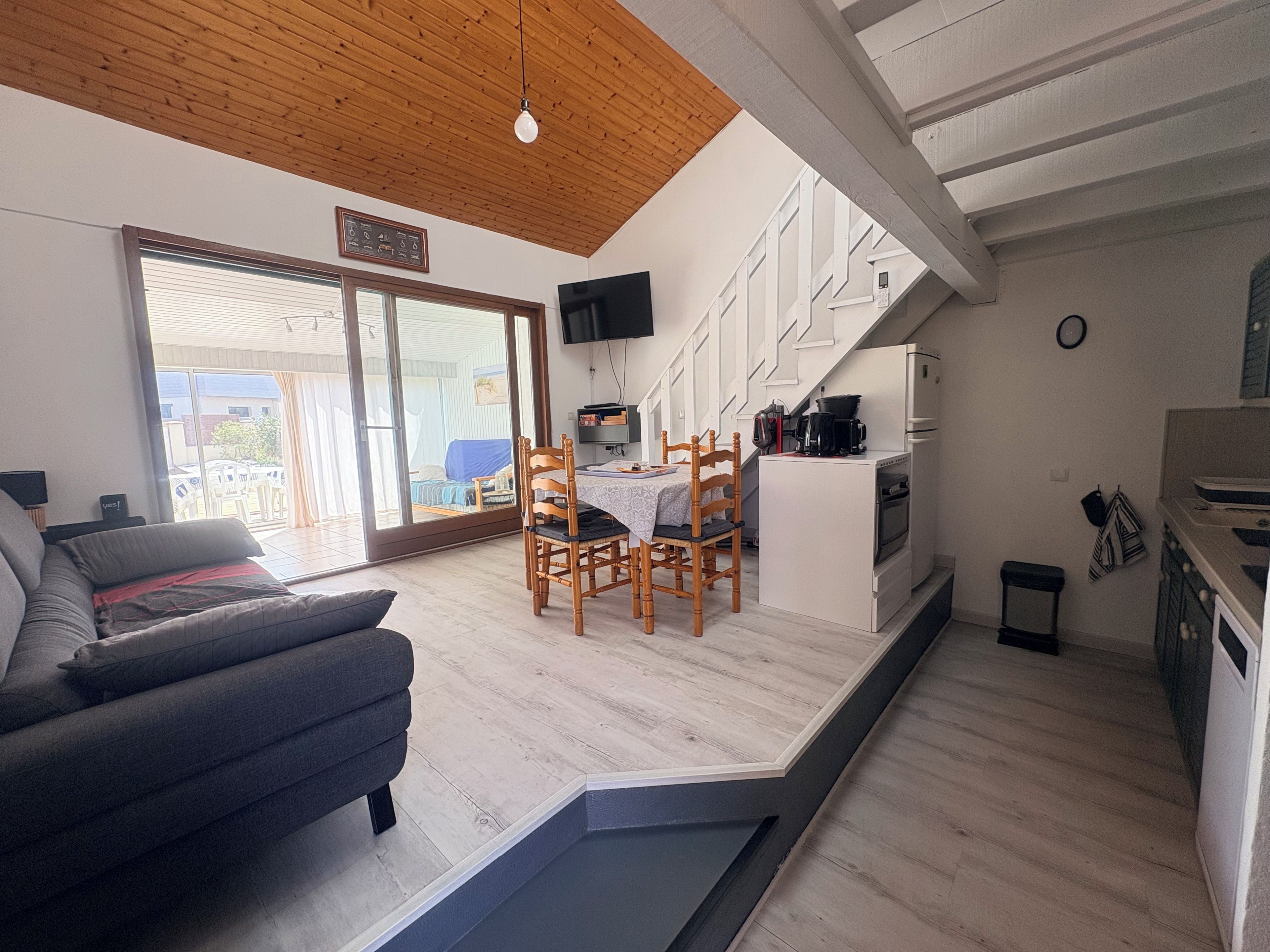 A modern living room with a sofa, a dining table with chairs, a kitchen area with appliances, and a large sliding glass door leading to an outdoor area.