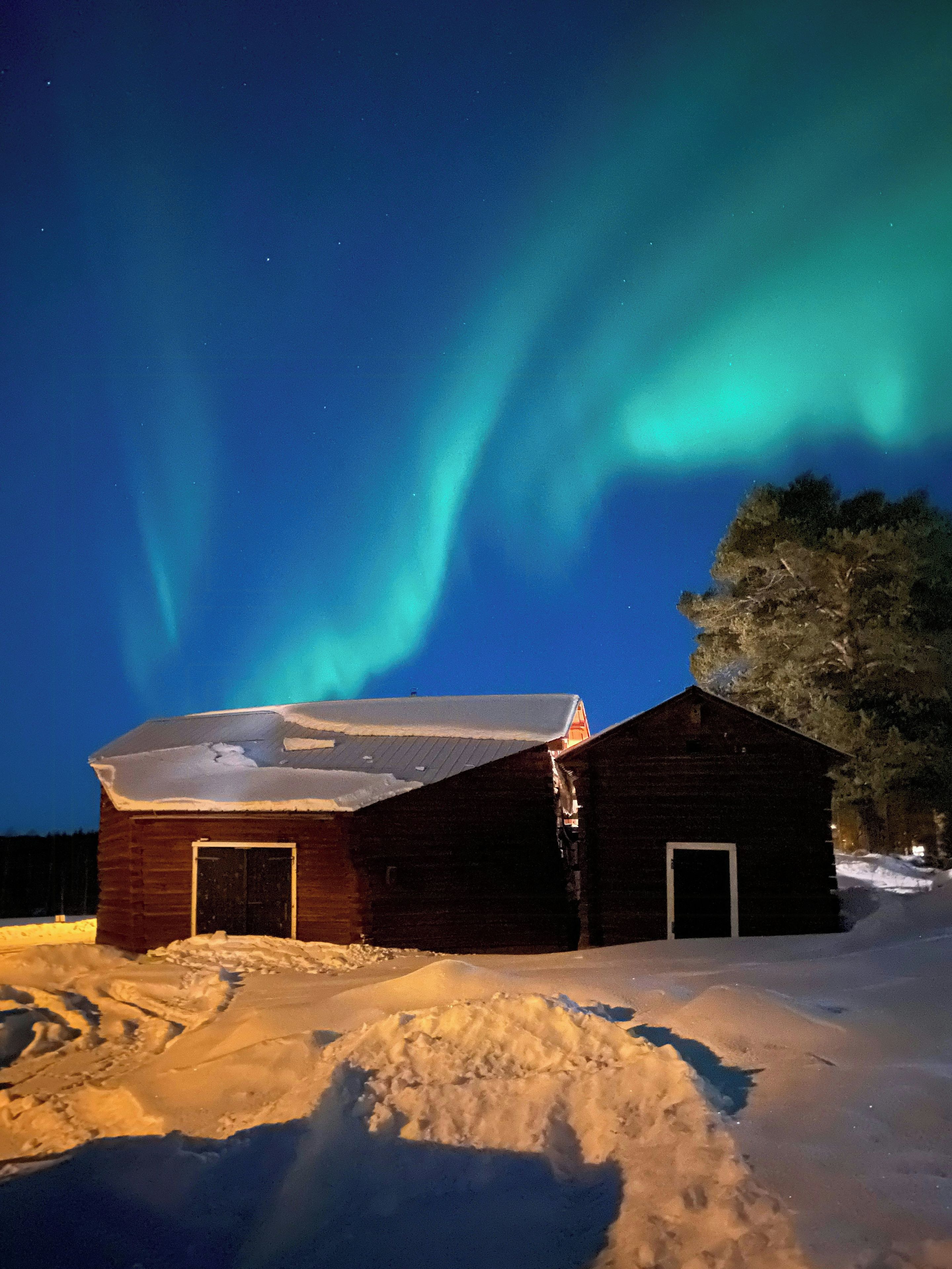 En træhytte med snebetægt tag og en snedrive foran, under en nattehimmel med nordlys.