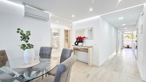 Een moderne woonkamer met een glazen eettafel, grijze gestoffeerde stoelen, een witte console met een rode plant in pot, en een grote spiegel aan de muur.