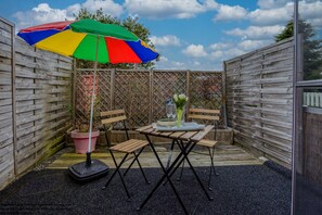 Eine Terrasse mit einem Holztisch, zwei Stühlen, einem bunten Sonnenschirm und einem Blumentopf.
