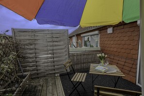 Eine Terrasse mit Holzlattenzaun, einem Tisch mit Buch und Gläsern sowie ein Fenster mit Blumen.