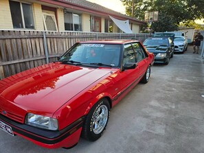 Une voiture rouge est garée dans une allée, avec une clôture en bois et un bâtiment en arrière-plan.