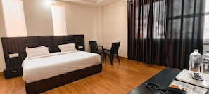Habitación de hotel con una cama grande, un escritorio, dos sillas, un ventanal con cortinas y vistas a la ciudad.