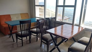 A dining area with a wooden table and chairs, a colorful sofa, and large windows with a view of mountains. - Eden Resort By Live Inn  (Lansdowne)