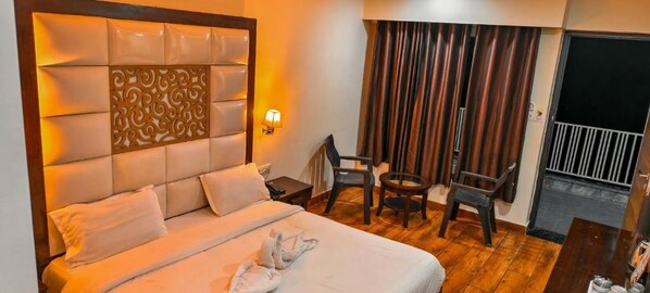 Habitación de hotel con una cama grande, un cabecero decorativo, un rincón de descanso con una silla y una mesita, y vistas al exterior a través de cortinas.