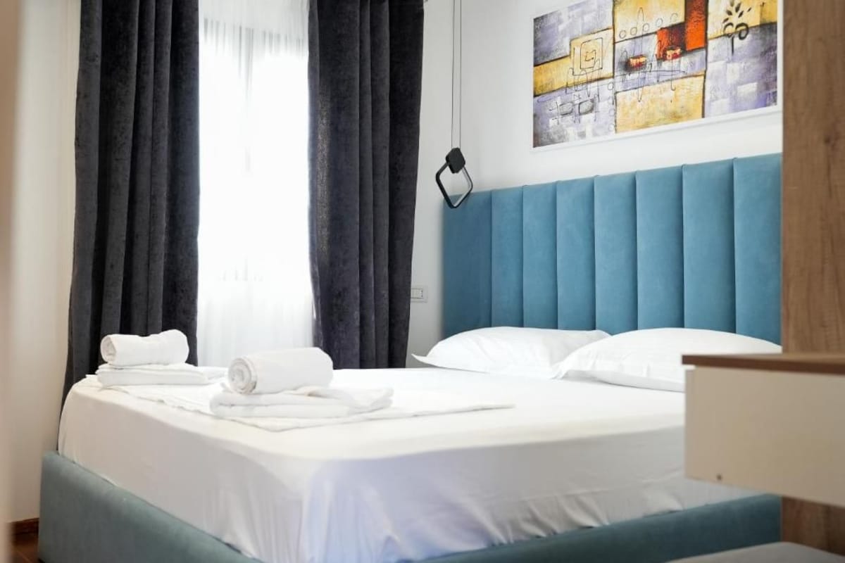 A hotel room with a bed, white linens, a blue headboard, and a painting on the wall.