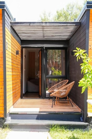 Un espacio exterior moderno con terraza de madera, puerta corrediza de vidrio que da a una habitación interior y dos sillas de mimbre.
