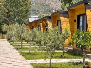 Una hilera de cabañas de madera con puertas numeradas, un camino pavimentado y árboles en un área ajardinada.