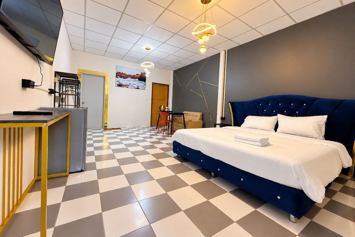Una habitación de hotel con piso a cuadros, una cama con cabecera azul, un escritorio con una silla, un televisor y un cuadro en la pared.