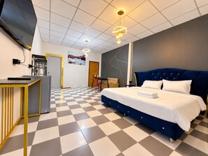 Una habitación de hotel con piso a cuadros, una cama con cabecera azul, un escritorio con una silla, un televisor y un cuadro en la pared.