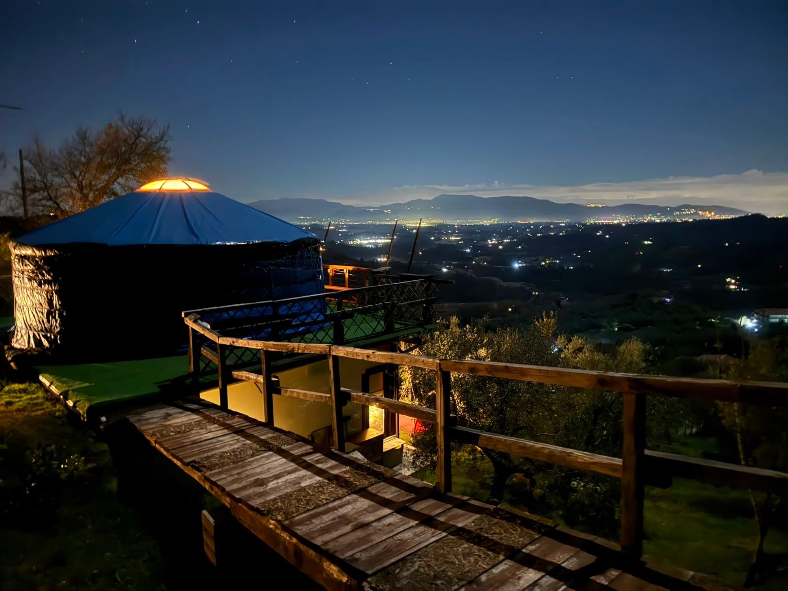 A yurt with a blue roof, a wooden deck, and a view of a cityscape at night.