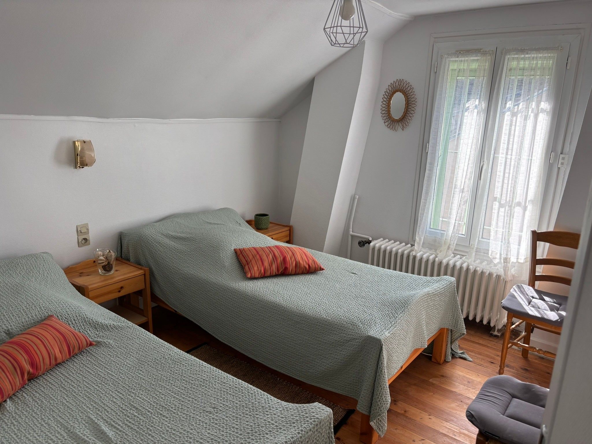 Une chambre avec deux lits, un radiateur, une fenêtre avec des rideaux, une chaise et une petite table avec une lampe.