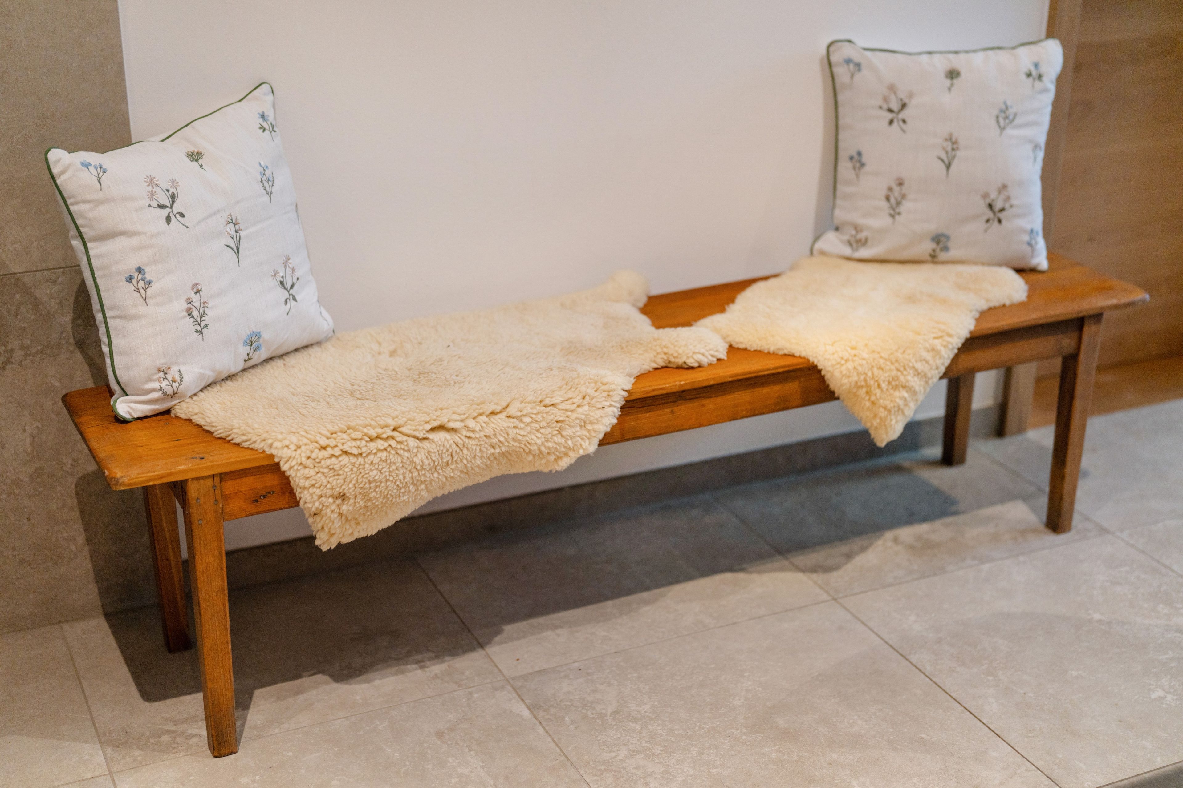 A wooden bench with a beige blanket and two pillows with floral embroidery.