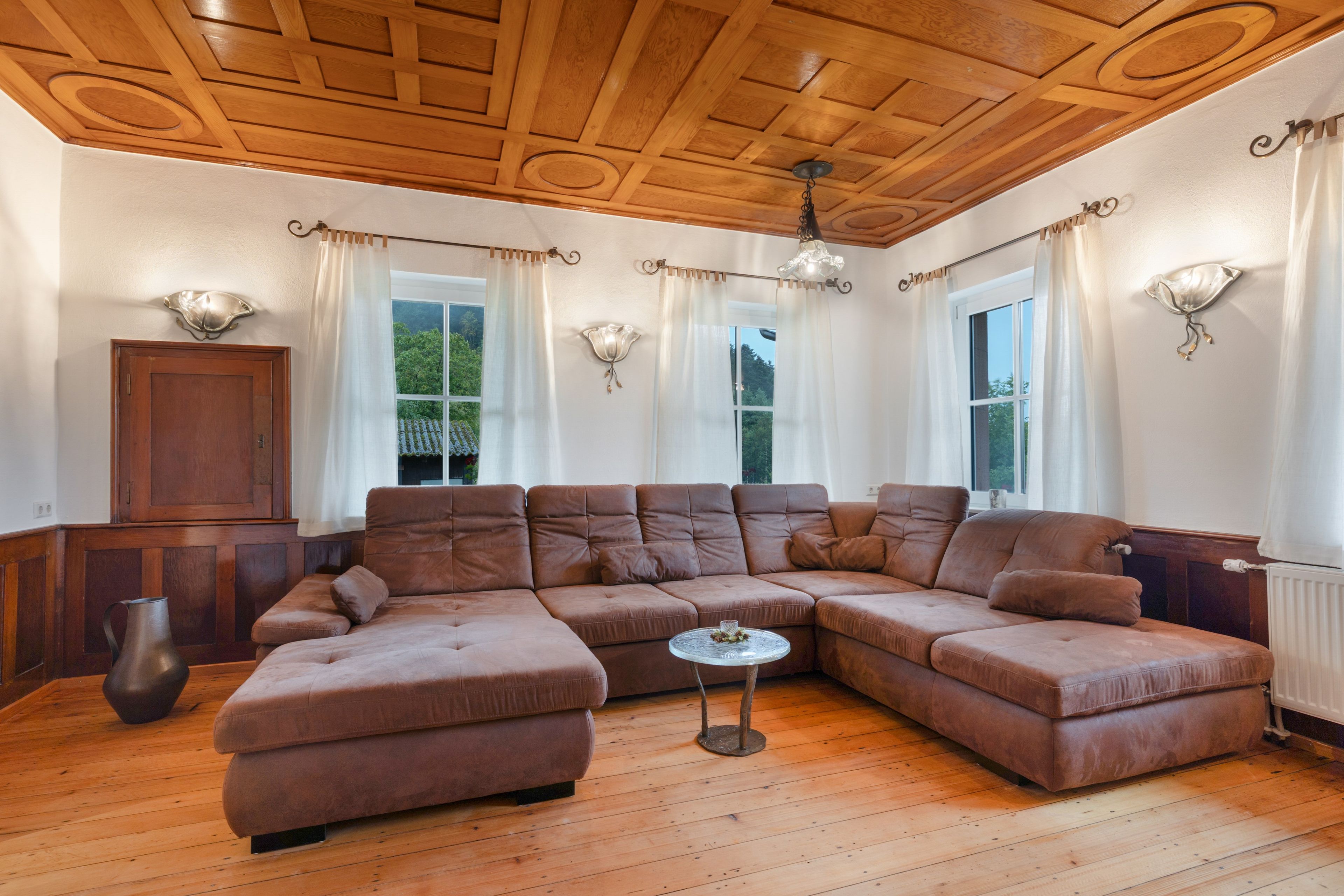 A spacious living room with a large L-shaped sectional sofa, wooden flooring, and a wooden ceiling with intricate designs.