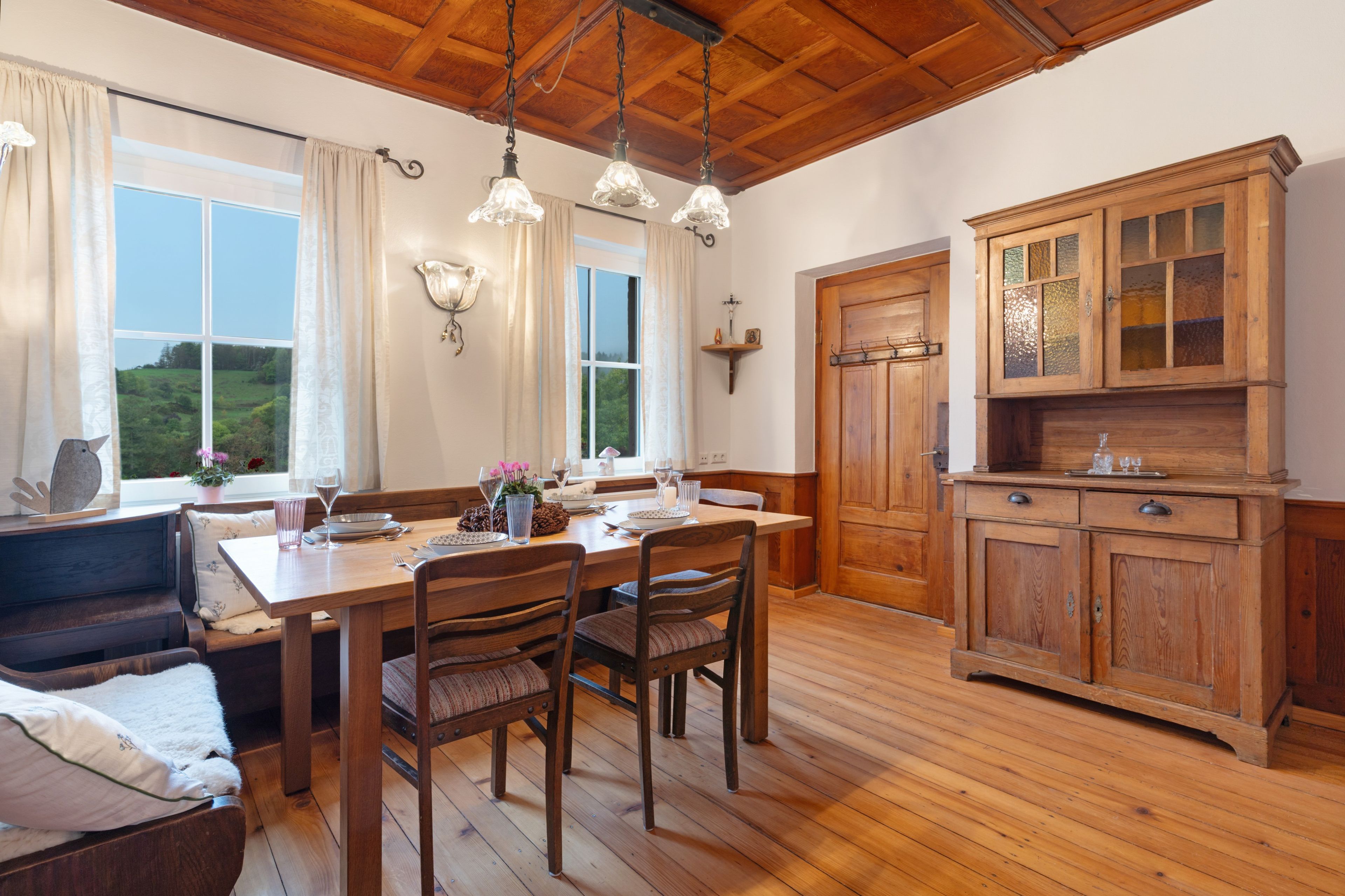 A dining area with a wooden table, chairs, and a bench. There are windows with curtains, a wooden cabinet, and a ceiling with wooden beams.
