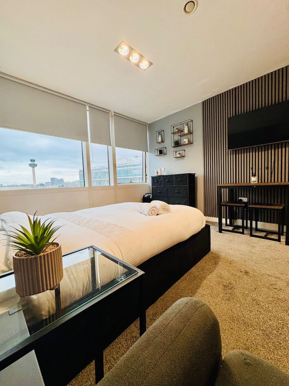 A modern hotel room with a large bed, a desk, a chair, a TV, and a city view.