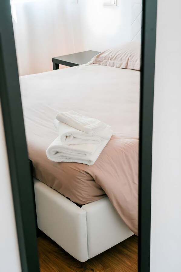 Un lit bien fait, avec une serviette blanche pliée, une table de chevet en bois et un luminaire fixé au mur.