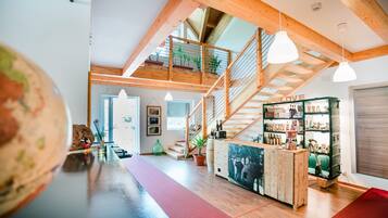 Une pièce spacieuse avec un plafond haut, des poutres en bois, un escalier, un coin bar avec des bouteilles et une grande fenêtre.