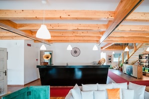 Un hall d'hôtel comprenant un comptoir d'accueil, des poutres en bois au plafond, des luminaires suspendus, une horloge murale et un escalier menant à un espace bar.