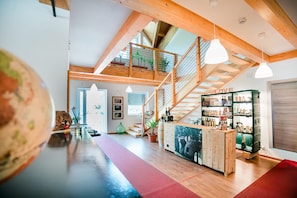 A spacious room with a high ceiling, wooden beams, a staircase, a bar area with bottles, and a large window. - Room 8 in Borgo Valsugana – double or triple, self check-in (Borgo Valsugana)