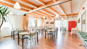 Uma sala de jantar ampla com várias mesas e cadeiras, vigas de madeira no teto e grandes janelas que permitem entrada de luz natural.