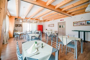 Uma sala de jantar com várias mesas e cadeiras, vigas de madeira no teto e quadros decorativos na parede.