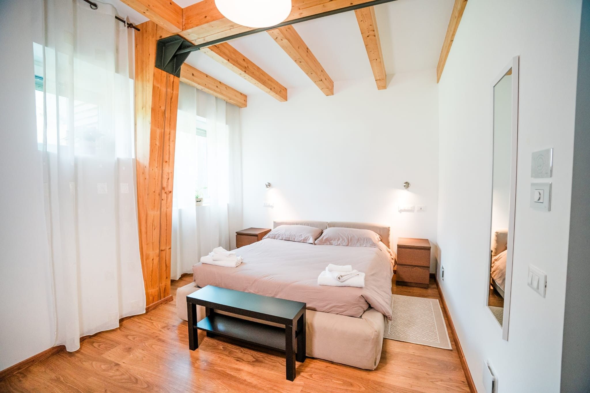 Une chambre à coucher comprenant un lit, des tables de chevet, un banc et un plafond avec des poutres apparentes.