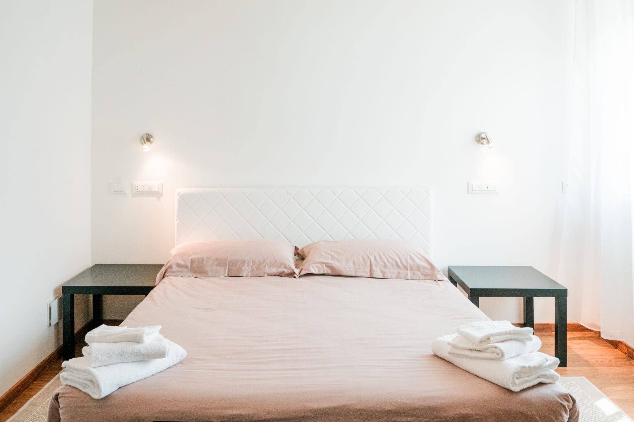 Un lit bien fait, avec une tête de lit matelassée, deux tables de chevet et une paire de serviettes posées sur le sol.