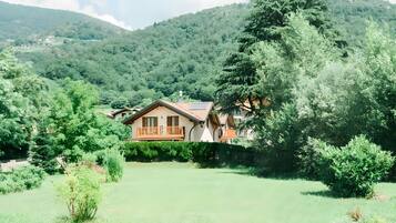 Une maison dotée d’un balcon, entourée d’un champ verdoyant et d’arbres, avec des montagnes en arrière-plan.