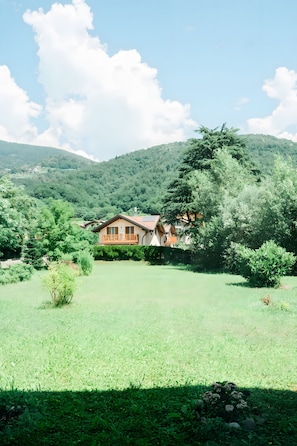 Una casa con balcón, rodeada de un campo cubierto de césped y árboles, con montañas al fondo.