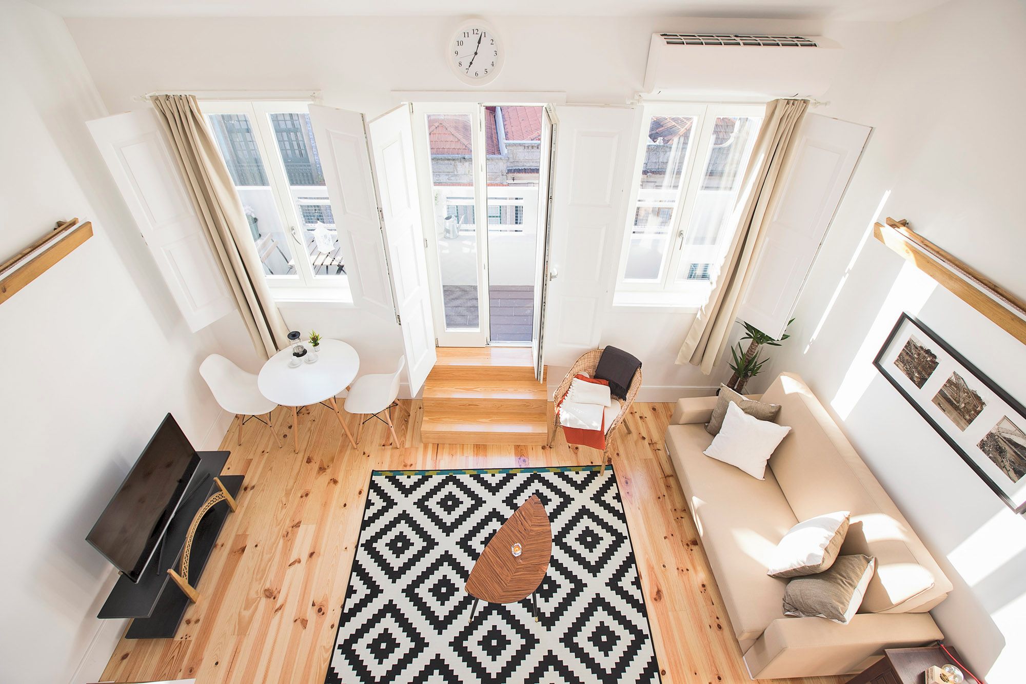 A modern living room with a sofa, a coffee table, and a TV. There are large windows with curtains, a patterned rug, and a clock on the wall.