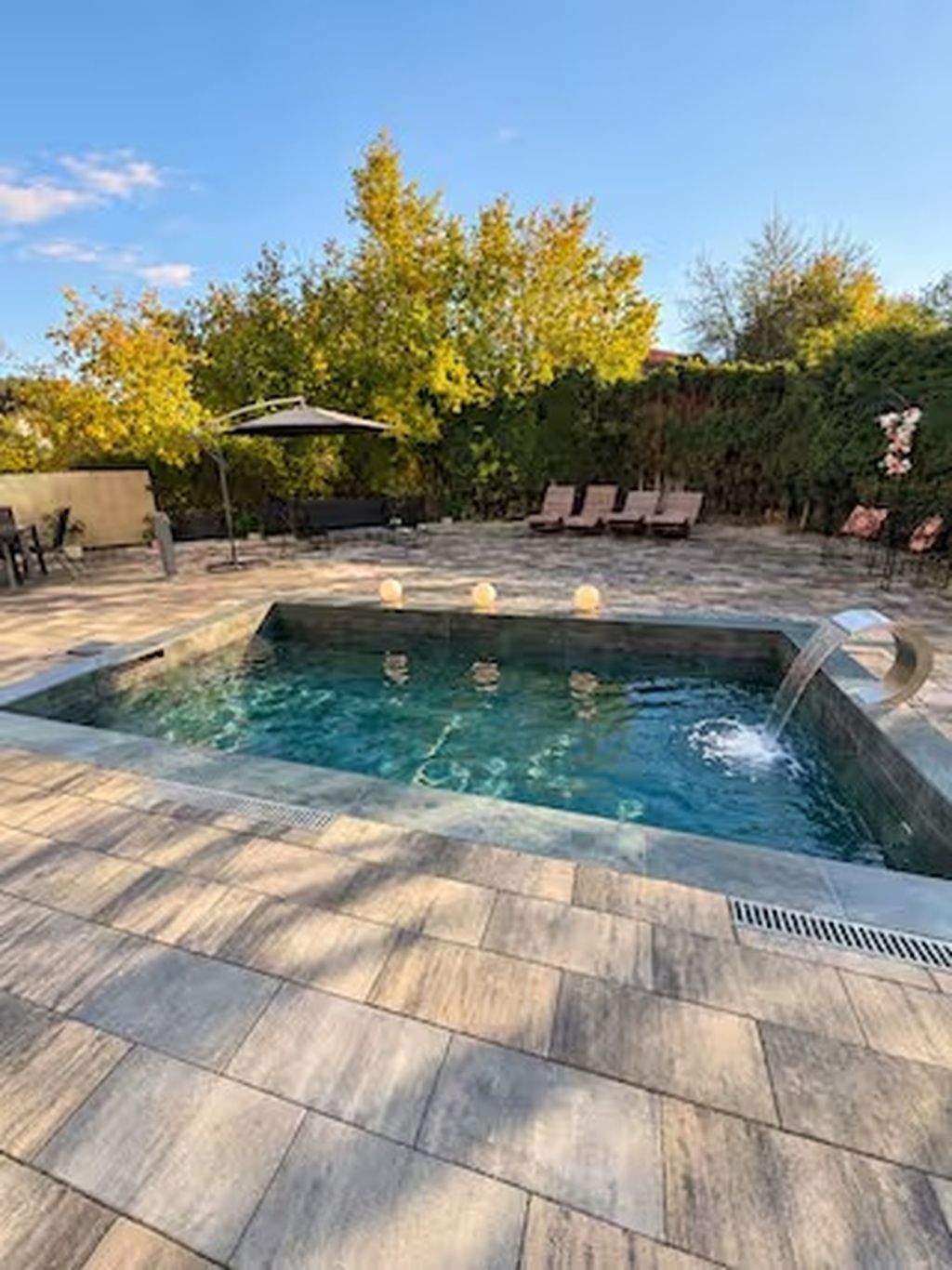 A rectangular swimming pool with a stone tile border, surrounded by a patio area with lounge chairs and a covered seating area.
