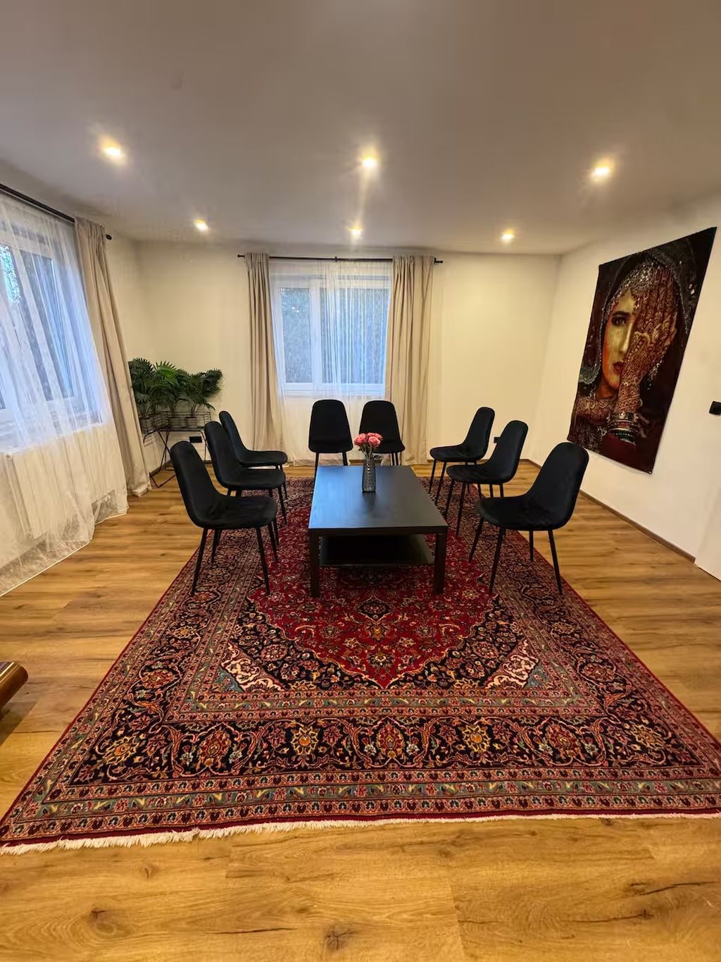 Ein Konferenzraum mit einem dunklen Holztisch, sechs schwarzen Stühlen, einem roten Teppich mit Muster und einem großen Fenster mit Vorhängen. An der Wand hängt ein Gemälde.