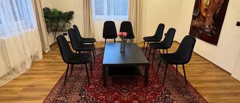 Ein Konferenzraum mit einem dunklen Holztisch, sechs schwarzen Stühlen, einem roten Teppich mit Muster und einem großen Fenster mit Vorhängen. An der Wand hängt ein Gemälde.
