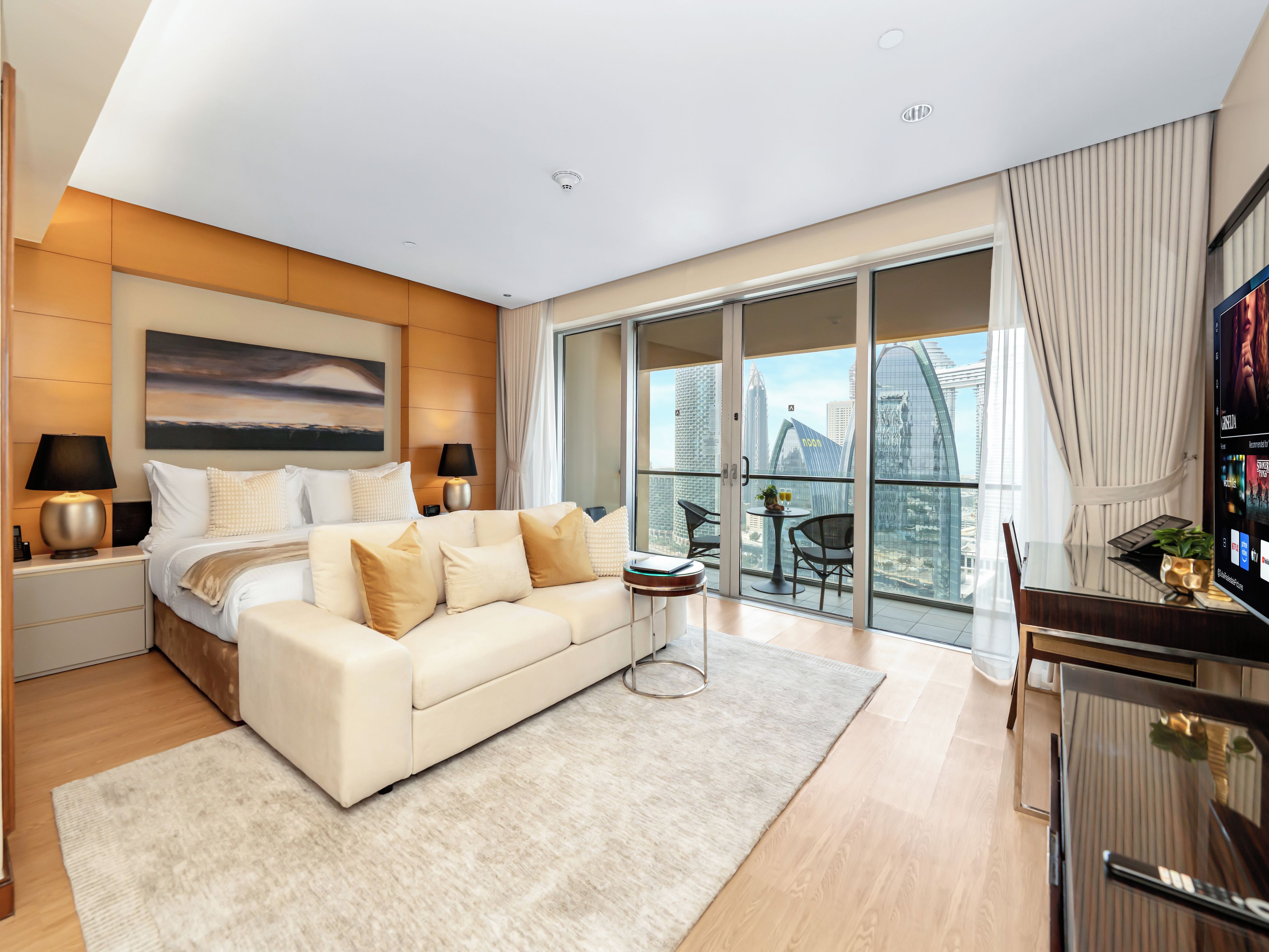 A modern hotel room with a large bed, a desk, a TV, and a balcony with a city view.