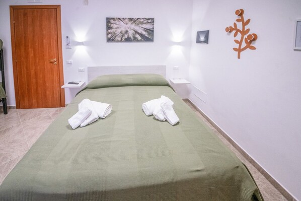 A hotel room with a bed, two bedside tables, a wooden door, a wall-mounted plant, and a painting.